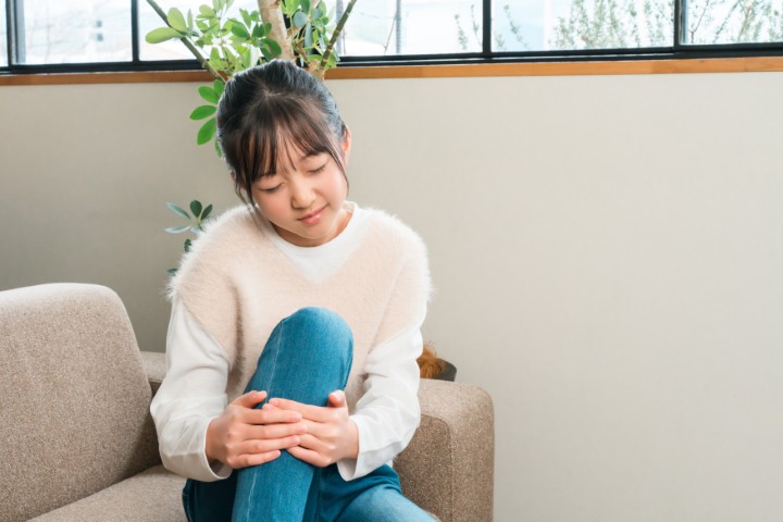 A young girl, either elementary or middle school age, is sitting on the sofa at home, clutching her knee and shin due to knee pain.
